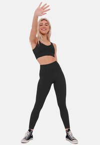 Black sports bra and high-waisted leggings, both form-fitting. Worn with black high-top sneakers. Model raises hand, smiling.