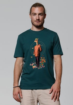 Cyaan t-shirt met een grafisch ontwerp van een giraffe in een bruin pak, omringd door bloemen en vlinders. Korte mouwen en een ronde halslijn.