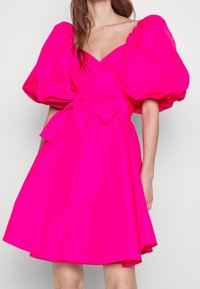 Vrouw in een heldere roze jurk met volumineuze pofmouwen, V-hals en een grote gestrikte strik op de taille, staand tegen een effen achtergrond.