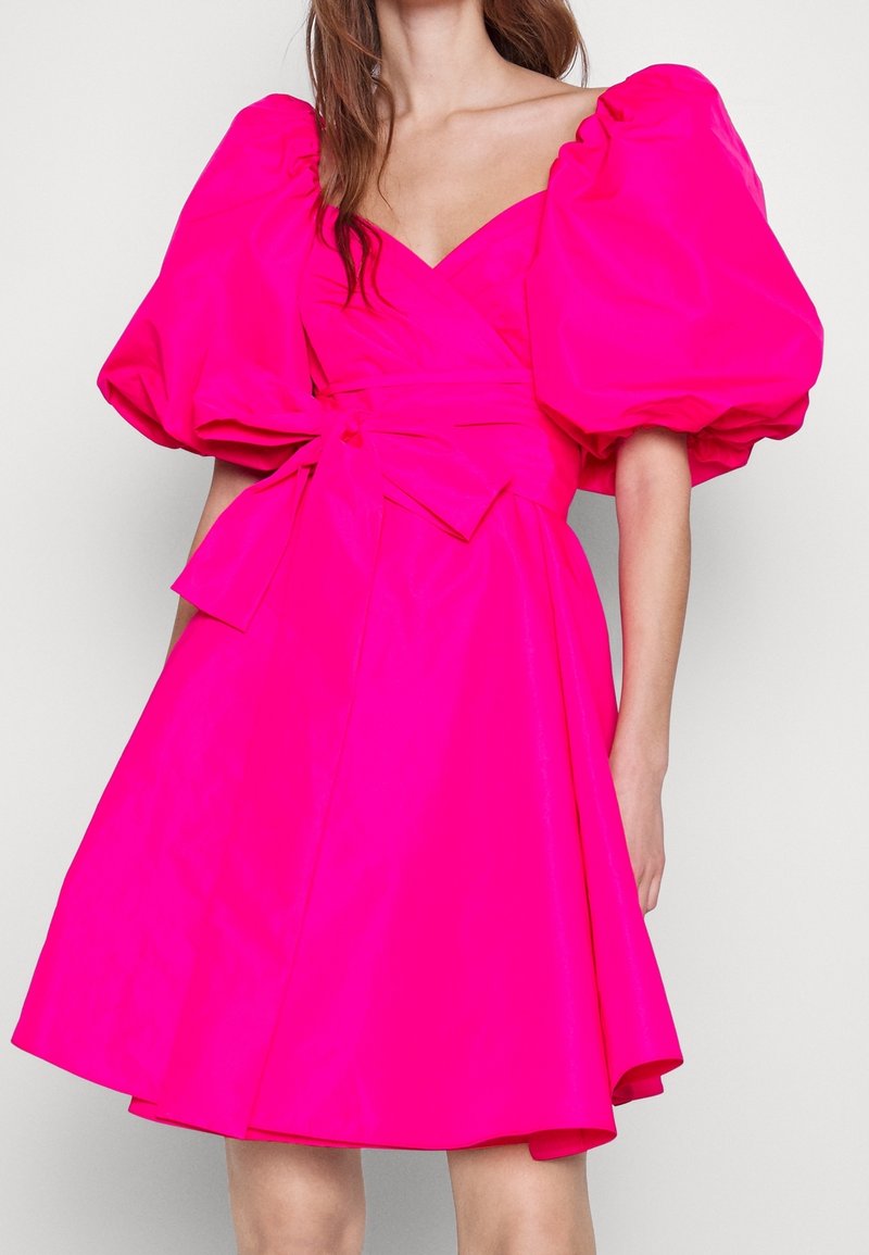 Vrouw in een heldere roze jurk met volumineuze pofmouwen, V-hals en een grote gestrikte strik op de taille, staand tegen een effen achtergrond.