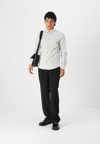 Lichtgrijze blouse, zwarte broek en glanzende zwarte schoenen. De persoon houdt een kleine zwarte tas vast en staat tegen een witte achtergrond.