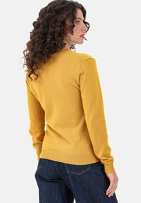 Gelber Strickpullover mit rundem Ausschnitt, langen Ärmeln und gerippten Bündchen. Kombiniert mit dunklen Denim-Jeans, die eine figurbetonte Silhouette zeigen.