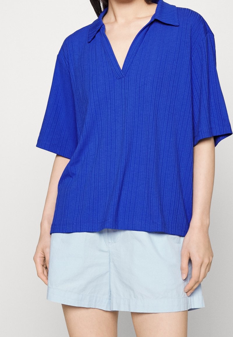 Femme portant un polo bleu vif à manches courtes côtelé avec un col en V et un short bleu clair, debout devant un fond uni.