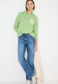 Vrouw in een groene hoodie en blauwe spijkerbroek leunt tegen een muur, houdt een beige fleecejack vast en draagt zilveren sneakers.