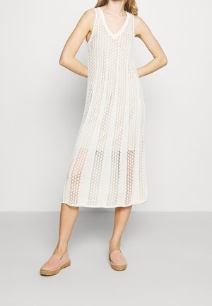 Femme portant une robe midi blanche en crochet sans manches avec un décolleté en V et des espadrilles roses, debout les mains sur les hanches.