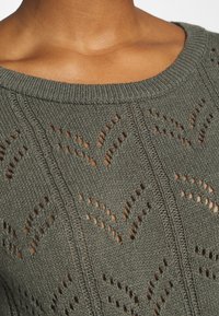 Pull en maille vert pâle avec un col rond, présentant un motif ajouré et un design subtil en zigzag le long du tissu.