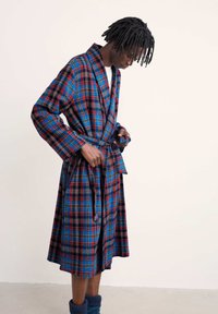 Karo bathrobe u plavoj, crvenoj i crnoj boji s dugim rukavima, remenom i džepovima; teksturirana tkanina, dužine do koljena. Model pozira dok veže remen.