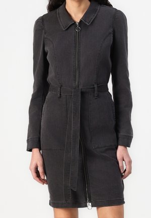 Donna che indossa un vestito lungo in denim nero a maniche lunghe, con cerniera frontale, colletto, due tasche frontali e una cintura in tessuto abbinata legata in vita.