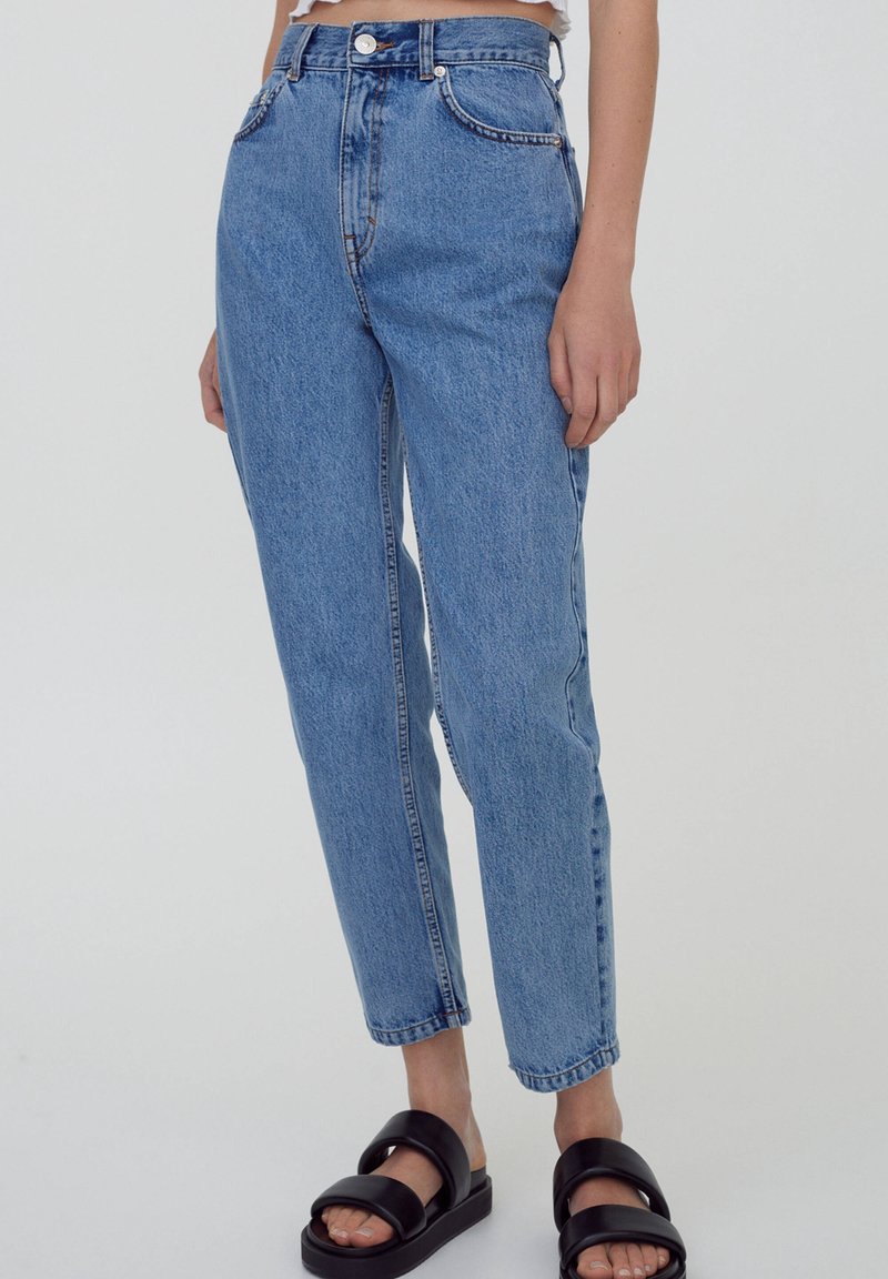 Jean bleu taille haute en denim, avec une coupe droite, des surpiqûres contrastées et des poches avant. Porté avec des sandales noires.