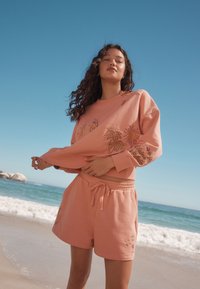 Completo felpa e pantaloncini rosa chiaro con ricami di sole e palme, materiale morbido, vestibilità rilassata e cintura regolabile.