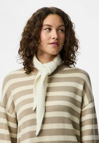 Ung kvinde med krøllet hår iført en beige og hvid stribet sweater og et blødt hvidt tørklæde bundet om halsen, kigger til siden.