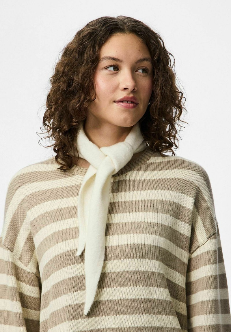 Ung kvinde med krøllet hår iført en beige og hvid stribet sweater og et blødt hvidt tørklæde bundet om halsen, kigger til siden.