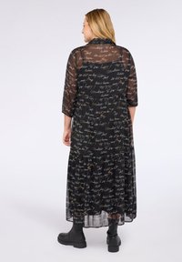Donna con capelli biondi che indossa un lungo vestito nero trasparente con testo bianco, stivali neri alla caviglia, in piedi su uno sfondo bianco uniforme.