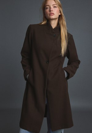 Femme aux longs cheveux blonds portant un manteau boutonné marron foncé, les mains dans les poches, sur un fond gris foncé uni.