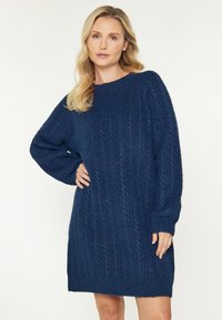 Blond kvinna står med handen på höften iförd en marinblå kabelstickad klänning mot en vit bakgrund.
