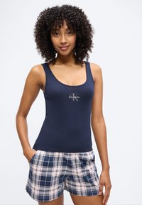 Νεαρή γυναίκα με σγουρά μαλλιά, φοράει μπλε σκούρο αμάνικο τοπ Calvin Klein και μπλε λευκά καρό σορτς, με τα χέρια στις τσέπες.