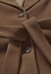 Brauner Wollmantel mit Reverskragen, Bindegürtel und Knopfverschluss. Der Stoff hat eine weiche Textur mit dezentem Glanz.
