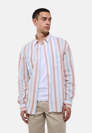 Homme portant une chemise à manches longues boutonnée avec des rayures verticales bleues, blanches et brunes sur un t-shirt blanc et un pantalon beige.