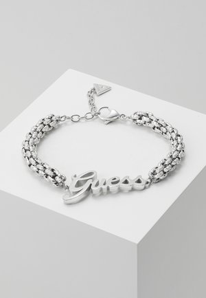 Srebrna pleciona bransoletka z łańcuszkiem, z centralnym elementem z logo "Guess" oraz małym trójkątnym zawieszonym na regulowanym zapięciu, prezentowana na białej powierzchni.