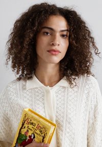 Cardigan alb din dantelă purtat peste un top deschis la culoare, având un guler. Ținând în mână o carte cu coperta galbenă "Albă ca Zăpada și cei șapte pitici".