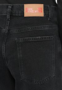 Jeansi negri din denim cu o buzunar pătrat pe spate și o etichetă maro cu "BAUM UND PFERDGARTEN" embosată în roz. Detalii de textură uzată vizibile.