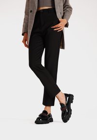 Pantaloni neri a vita alta con una vestibilità slim abbinati a mocassini neri lucidi decorati con gemme rosse. L'ensemble include un blazer a scacchi.