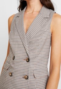 Rutig ärmlös blazer med djup V-ringning, kontrasterande rutor i beige och brunt, med dubbla knappar och skräddarsydd passform.