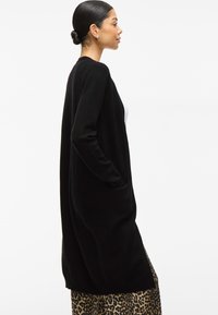 Svart, lång kofta i mjukt tyg, med öppen framdesign, ribbade muddar och sidofickor. Bärs över en mönstrad kjol.