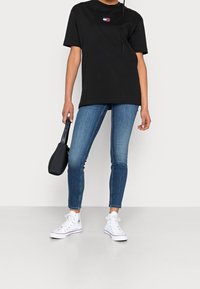 T-shirt en coton noir à manches courtes avec un petit logo, associé à un jean skinny bleu et des baskets montantes blanches. Sac à main noir.