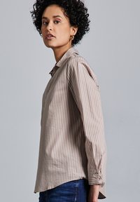 Camicia a maniche lunghe a righe marrone chiaro con una consistenza morbida e polsini abbottonati, indossata con jeans blu scuro su uno sfondo neutro.