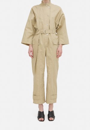 Beżowy bawełniany kombinezon z wysokim kołnierzem, dużymi rękawami, zapięciem na guziki z przodu, paskiem w talii i dwoma przednimi kieszeniami; noszony z czarnymi sandałami na obcasie.