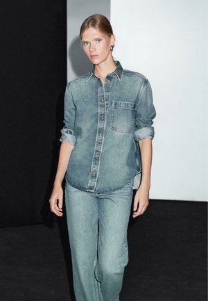 Vrouw met blond haar, gekleed in een lichtdenim shirt met opgerolde mouwen en bijpassende denim jeans, staande voor een zwart-witte achtergrond.