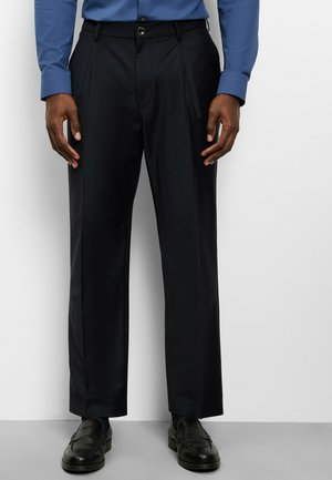 Man draagt zwarte rechtgesneden broek, zwarte loafers en een ingestoken blauw overhemd met lange mouwen, staand tegen een witte muur.