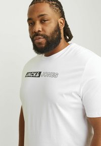 Camiseta blanca de algodón con un gráfico negro de "JACK & JONES" en el pecho. La tela parece suave y ligera.