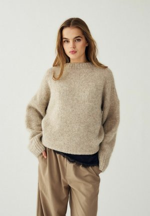 Mladá žena s dlhými vlasmi, oblečená v béžovom chlpatom svetri cez čierne čipkované tričko a vo voľných khaki nohaviciach, stojaca s rukami v vreckách.