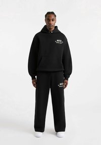 Completo felpa con cappuccio e pantaloni della tuta realizzato in tessuto morbido. Dotato di tasca frontale, dettagli con logo bianco e vestibilità comoda. Sneakers bianche completano l'outfit.