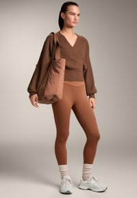 Barna kötött pulóver voluminózus ujjal, barna leggingssel párosítva. A modellen egy illeszkedő táska látható, és szürke sportcipőt visel.