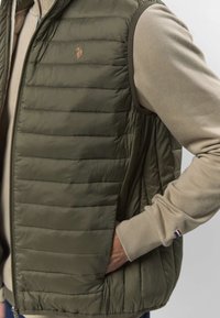 Bărbat purtând o vestă captusită verde măsliniu peste o cămașă bej cu mâneci lungi, cu mâna în buzunarul vestei, pe un fundal simplu.