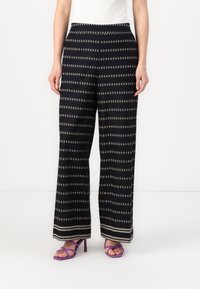 Μαύρα παντελόνια με φαρδιά μπατζάκια και χρυσά γεωμετρικά σχέδια, με επίπεδη μέση και στρίφωμα που τονίζεται από μια ελαφριά αντίθεση.