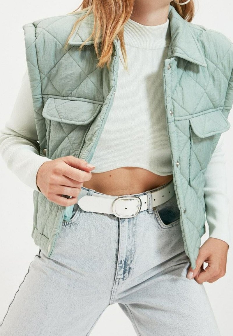 Jasnozielona pikowana kamizelka z dwoma przednimi kieszeniami, noszona na dopasowanym, jasnym swetrze. Połączona z spodami w kolorze jasnoniebieskim o wysokim stanie i białym paskiem.