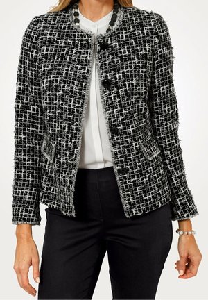 Kvinde iført sort-hvid tekstureret tweedjakke over hvid bluse, kombineret med sorte bukser og perlebesat halskæde og armbånd.