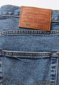 Pantalones vaqueros de mezclilla con un lavado azul claro, que presentan un parche de cuero marrón en relieve en la cintura etiquetado como "Levi Strauss & Co. Lot 501 W32 L."
