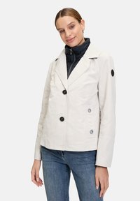 Light beige double-breasted jacket with black buttons, a notched collar, and a textured finish. Worn over a dark high-necked inner layer.