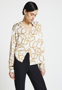 Camicia bianca con motivo a catena dorata. Maniche lunghe con polsini abbottonati, vestibilità comoda e scollo a V. Abbinata a pantaloni neri.