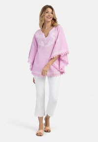 Lyserød bomulds-tunika med broderede blomstertryk, spidsdækkende kant og V-udskæring. Kombineret med hvide cropped bukser og lyserøde slidssandaler.