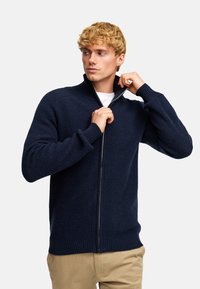 Granatowy sweter z suwakiem, prążkowany, z wysokim kołnierzem i długimi rękawami, wykonany z teksturowanej dzianiny, noszony częściowo rozpięty.