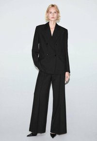 Czarna dwurzędowa marynarka z szerokimi klapami i dwoma przednimi kieszeniami, zestawiona z dopasowanymi spodniami o szerokich nogawkach oraz czarnymi, spiczastymi szpilkami.