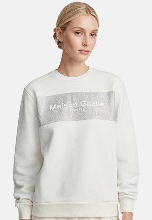 Mujer rubia con el cabello recogido en un moño bajo, vistiendo una sudadera blanca con el logo "Maison Gaelle Paris" de lentejuelas plateadas en el frente sobre fondo blanco.