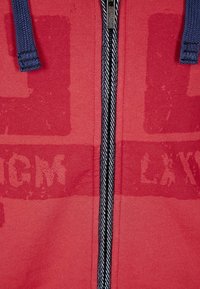 Sudadera con capucha roja con cremallera, hecha de tela suave, con una cremallera a rayas negra y blanca, y un patrón de texto blanco tenue en el frente.