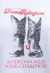 T-shirt blanc avec un graphique de deux bottes de cowboy noires ornées de détails complexes, accompagné de texte rouge : "Véritable Religion" et "Champion du Monde de Rodéo."